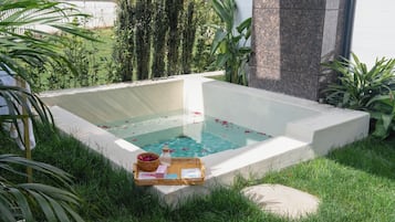 A square hot tub with a white interior, surrounded by lush greenery and a white fence in the background.