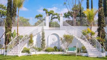 A white staircase leading to a gated entrance, surrounded by lush greenery and tall trees.
