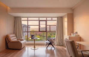 Habitació moderna amb un gran finestral, un sofà, una cadira, una taula petita i un escriptori amb una làmpada.