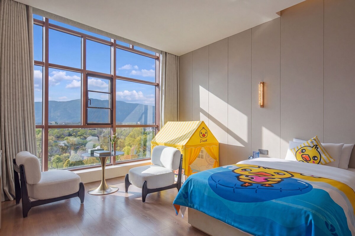 Ein Hotelzimmer mit großem Fenster, einem Bett mit einer Bettdecke mit einem Cartoon-Entenmuster, einem kleinen Tisch mit einer Vase und einem Stuhl.