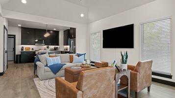 A modern living room with a sofa, coffee table, and chairs, a kitchen in the background, and a flat-screen TV mounted on the wall.