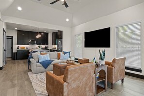 A modern living room with a sofa, coffee table, and chairs, a kitchen in the background, and a flat-screen TV mounted on the wall. - Hard Rock Hideaway @ Blue Whale/Route 66 (Catoosa)