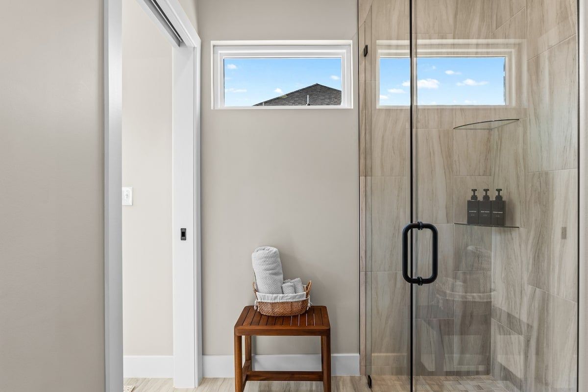 A modern bathroom with a glass shower enclosure, a small wooden table with towels, and a window with a view of a house.