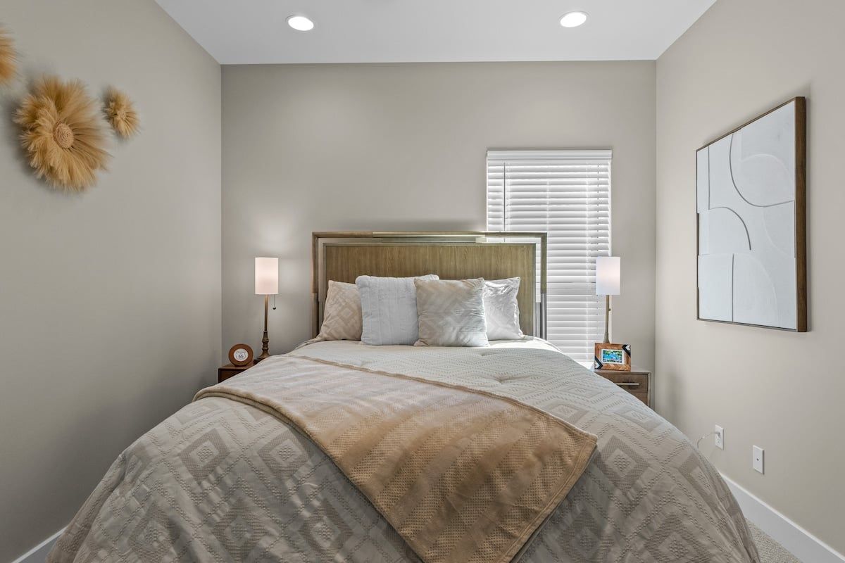 A bedroom with a large bed, two bedside tables, a window with blinds, and wall art.