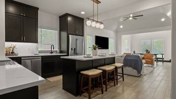 A modern kitchen with dark cabinetry, a central island with stools, a built-in refrigerator, and a ceiling fan.