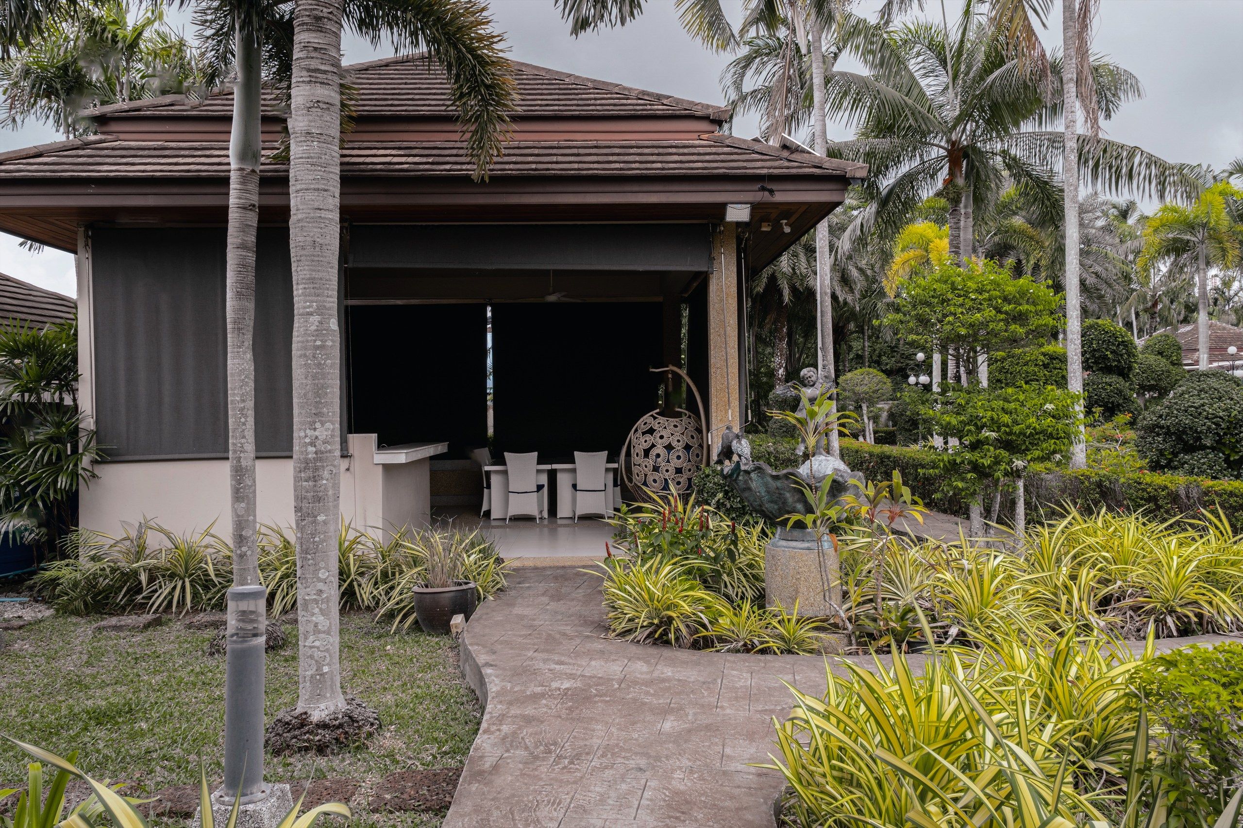 A covered outdoor seating area with a pathway leading to it, surrounded by tropical plants and palm trees.