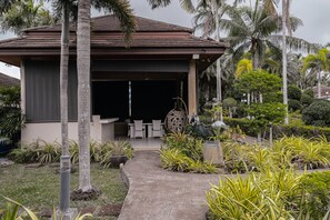 A covered outdoor seating area with a pathway leading to it, surrounded by tropical plants and palm trees. - The One Villa  (Mueang)