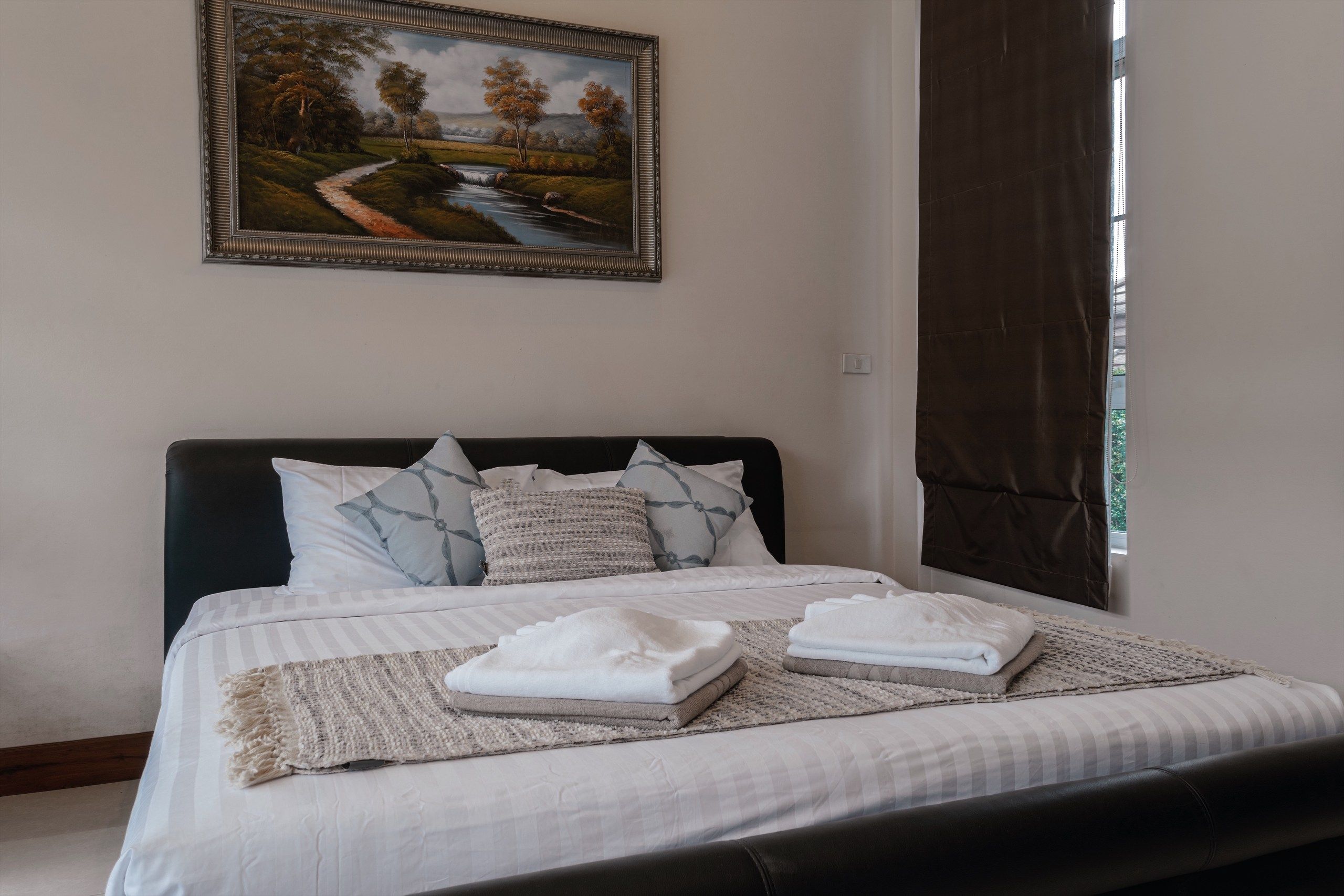 A neatly made bed with a white comforter and pillows, a painting of a landscape on the wall, and a window with curtains.