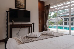 A hotel room with a bed, a television, a window with a view of a swimming pool and palm trees. - The One Villa  (Mueang)