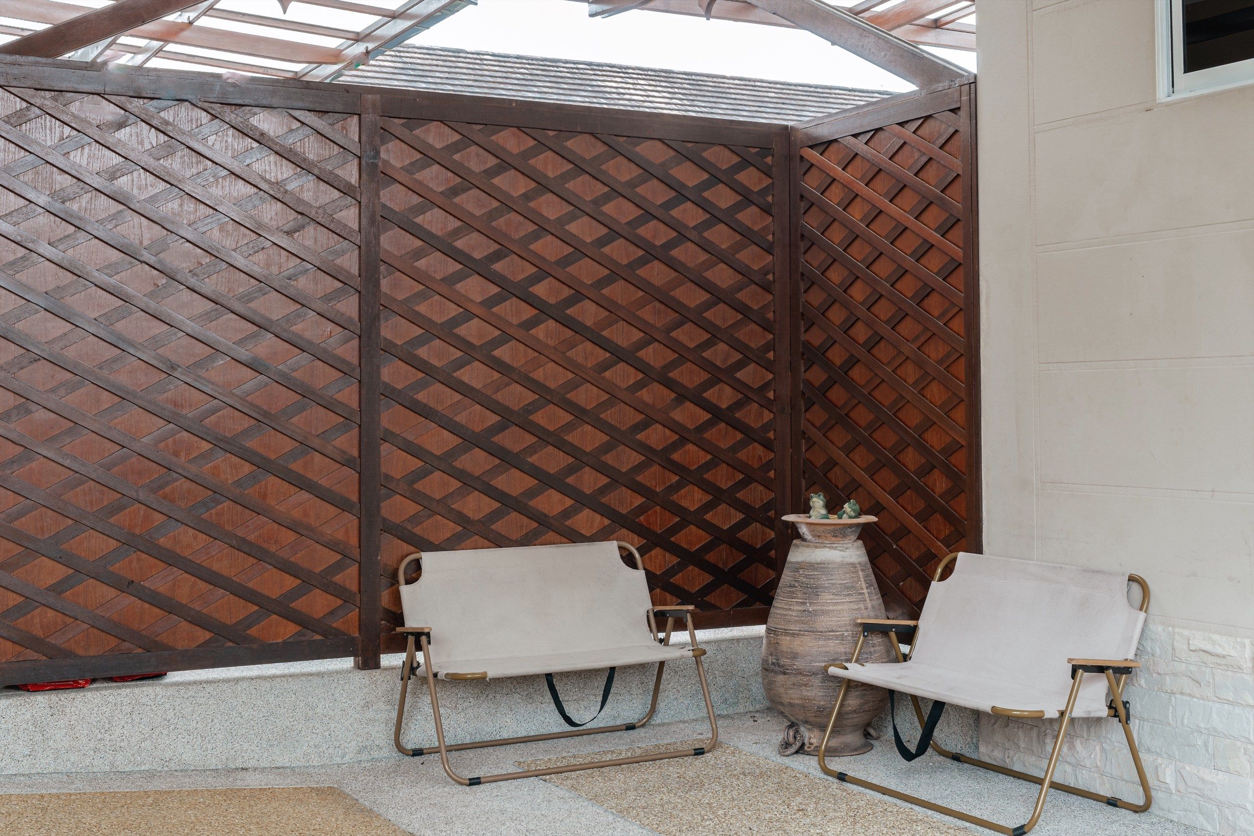 A covered outdoor seating area with two chairs and a table, a wooden lattice partition, and a stone planter.