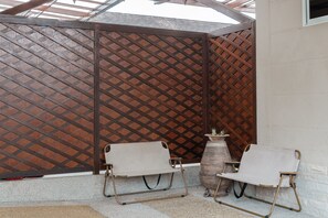 A covered outdoor seating area with two chairs and a table, a wooden lattice partition, and a stone planter.