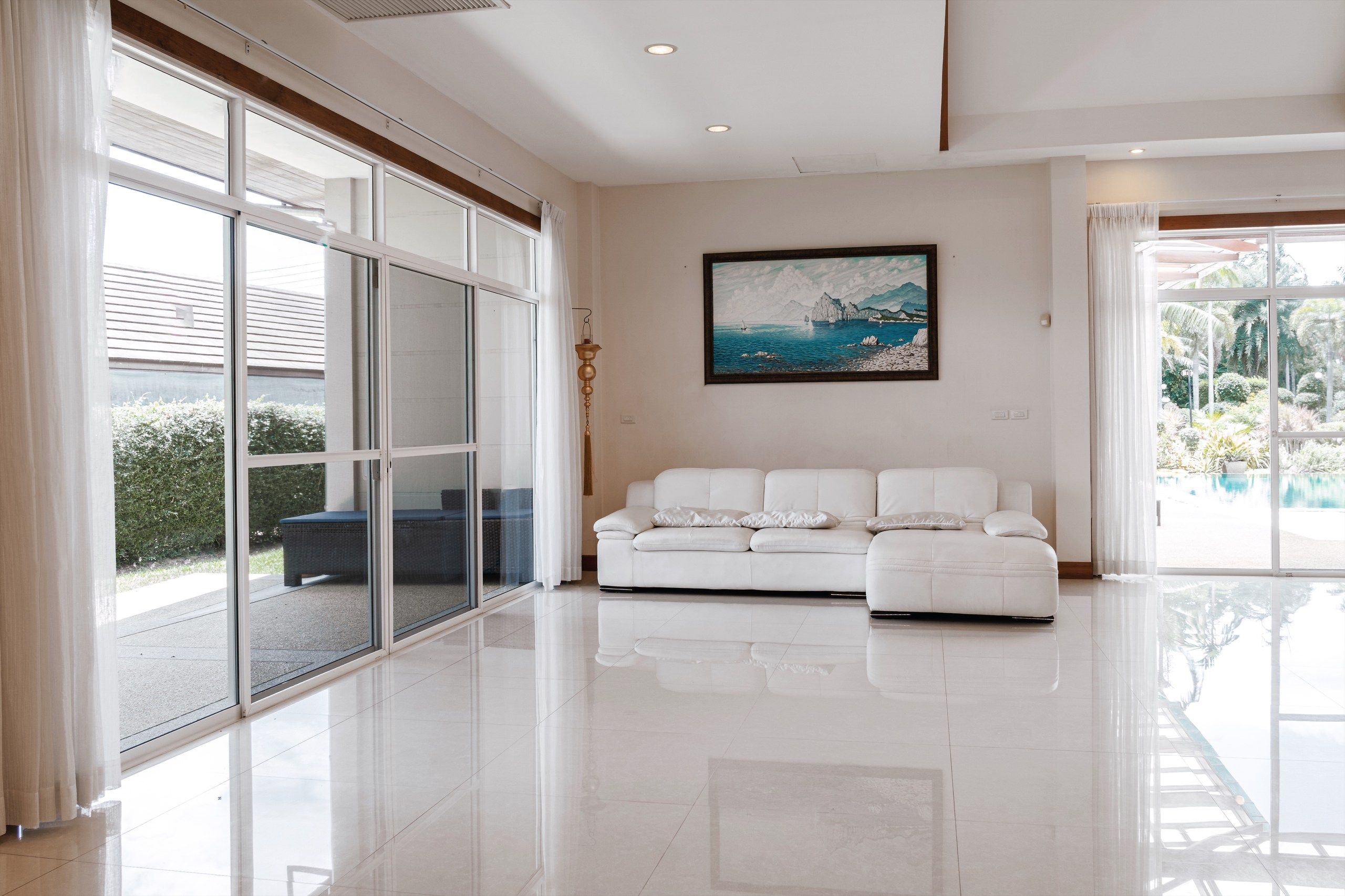 A spacious room with a white leather sofa, large sliding glass doors leading to a balcony, and a painting of a coastal landscape on the wall.