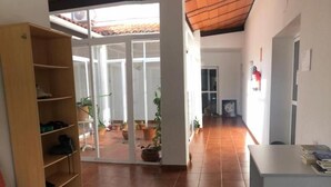 Um espaço interior com piso de azulejos, uma estante de madeira, uma mesa branca com livros e uma cadeira vermelha.