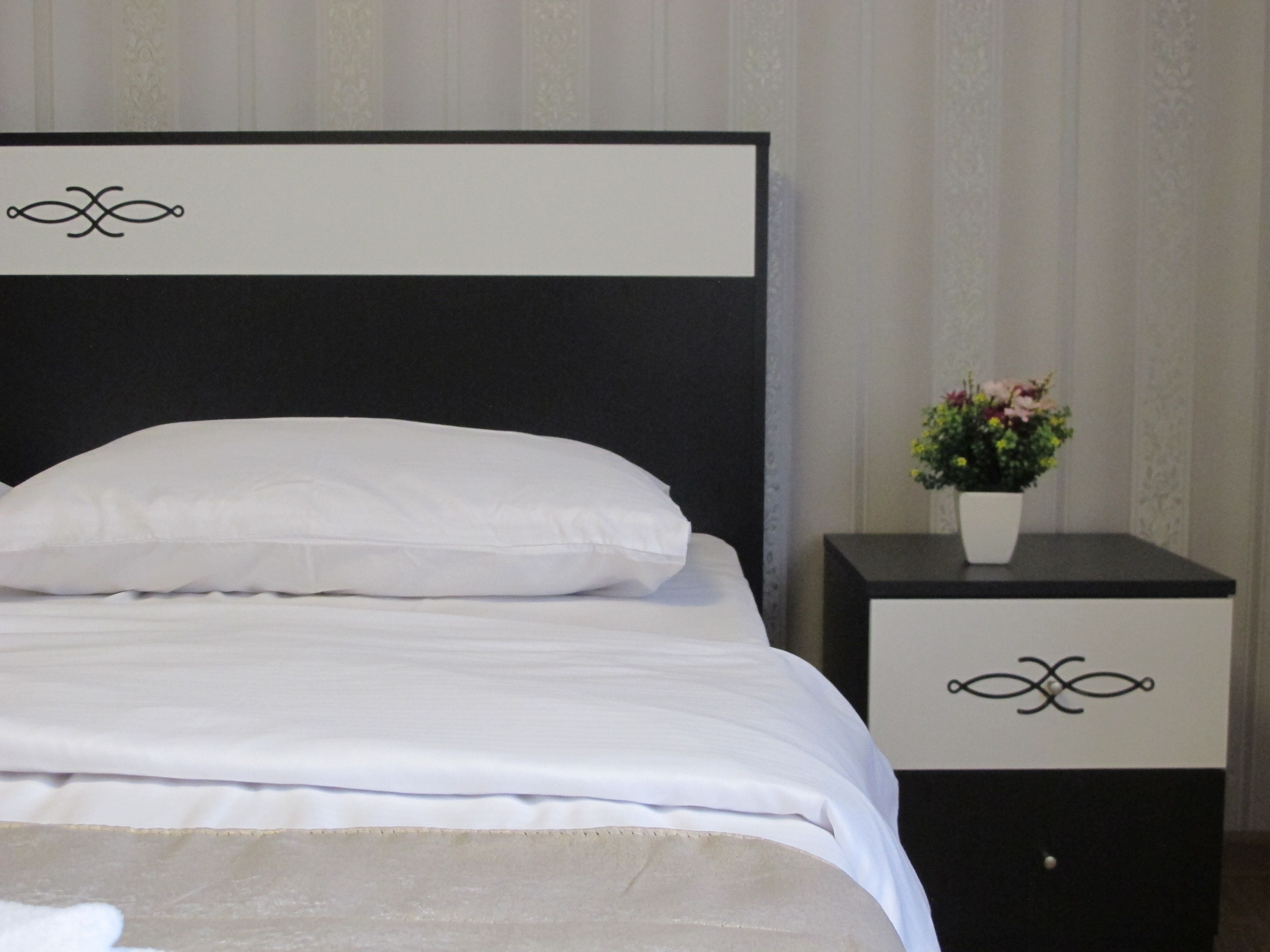 Una cama con ropa de cama blanca y cabecera negra, una mesita de noche con un florero, y un panel decorativo sobre la cama.