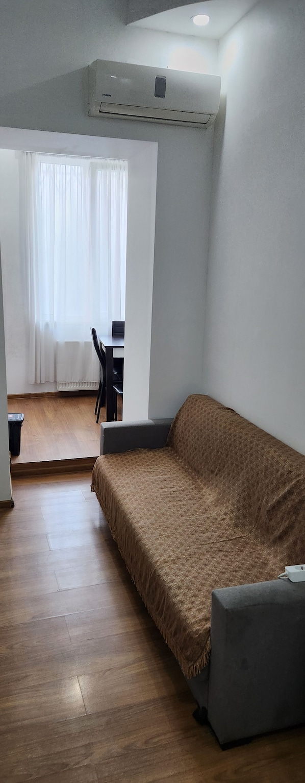 A room with a sofa, a table, and a chair. There is a window with curtains, a radiator, and an air conditioning unit on the wall. - Old Georgia (Tbilisi)