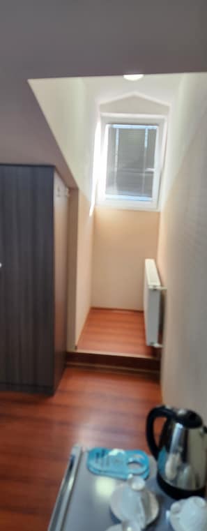 A small, narrow room with a sloped ceiling, a window with blinds, a radiator, and a kitchenette area with a kettle and cups. - Old Georgia (Tbilisi)