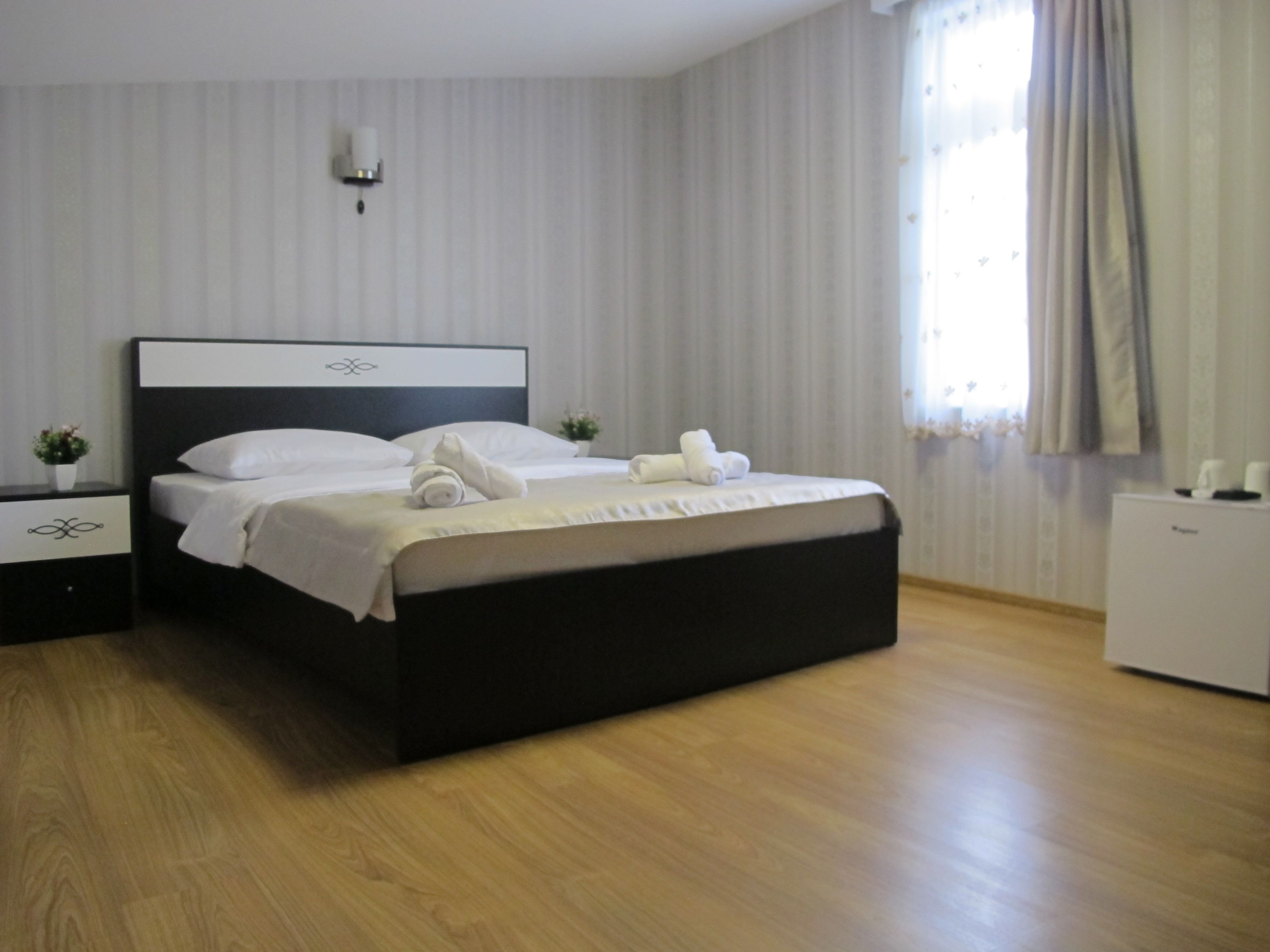 A hotel room with a bed, bedside tables, a nightstand, a dresser, a window with curtains, and a wall-mounted light fixture.