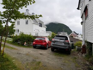 Un camino de grava con dos coches aparcados, una casa blanca con porche y una montaña al fondo.
