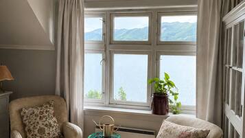 A room with two armchairs, a small table with a kettle, and a potted plant by a window with a view of mountains.