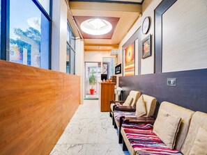 A hotel lobby with a reception desk, a waiting area with cushioned seating, and a view of the outdoors through large windows. - Deluxe JLN Marg Malviya Nagar Braj Haveli (Jaipur)