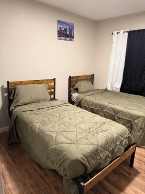 A room with two single beds with wooden frames and green bedding, a wall-mounted picture, and a black curtain. - Awesome 3-bedroom house with AC in vibrant Houston (Houston)