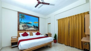 Ein Hotelzimmer mit einem Bett, einem Deckenventilator, einem Strandbild, einem Nachttisch, einer Topfpflanze und einem Fenster mit Vorhängen.