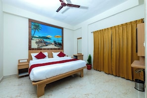 Quarto de hotel com cama, ventoinha no teto, pintura de uma praia, mesa de cabeceira, planta em vaso e janela com cortinas.
