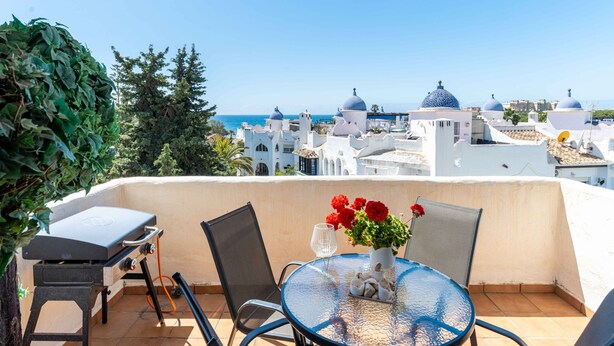 Eine Terrasse mit Tisch, Stühlen und Grill, mit Blick auf eine Küstenstadt mit weißen Gebäuden und blauen Kuppeln.