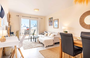 Ein modernes Wohnzimmer mit einer Couch, einem Esstisch und Stühlen. Ein großes Fenster bietet Blick auf Gebäude und eine Kuppelkonstruktion.