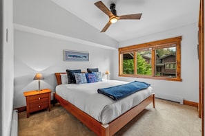 Quarto com cama, mesa de cabeceira, abajur, ventoinha no teto, janela com vista para o verde e quadro na parede.