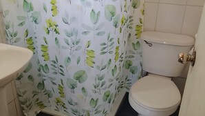 Une salle de bain avec des toilettes, un lavabo et un rideau de douche orné d’un motif de feuilles. Des tapis à rayures sont posés sur le sol.