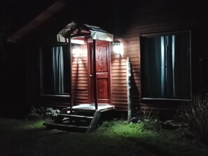 Een houten gebouw met een veranda en een deur, verlicht door buitenverlichting.
