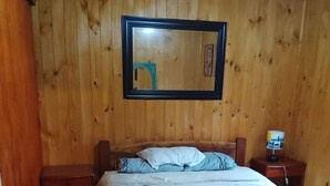 Une pièce aux murs lambrissés, contenant un lit, une table de chevet, une lampe et une porte.