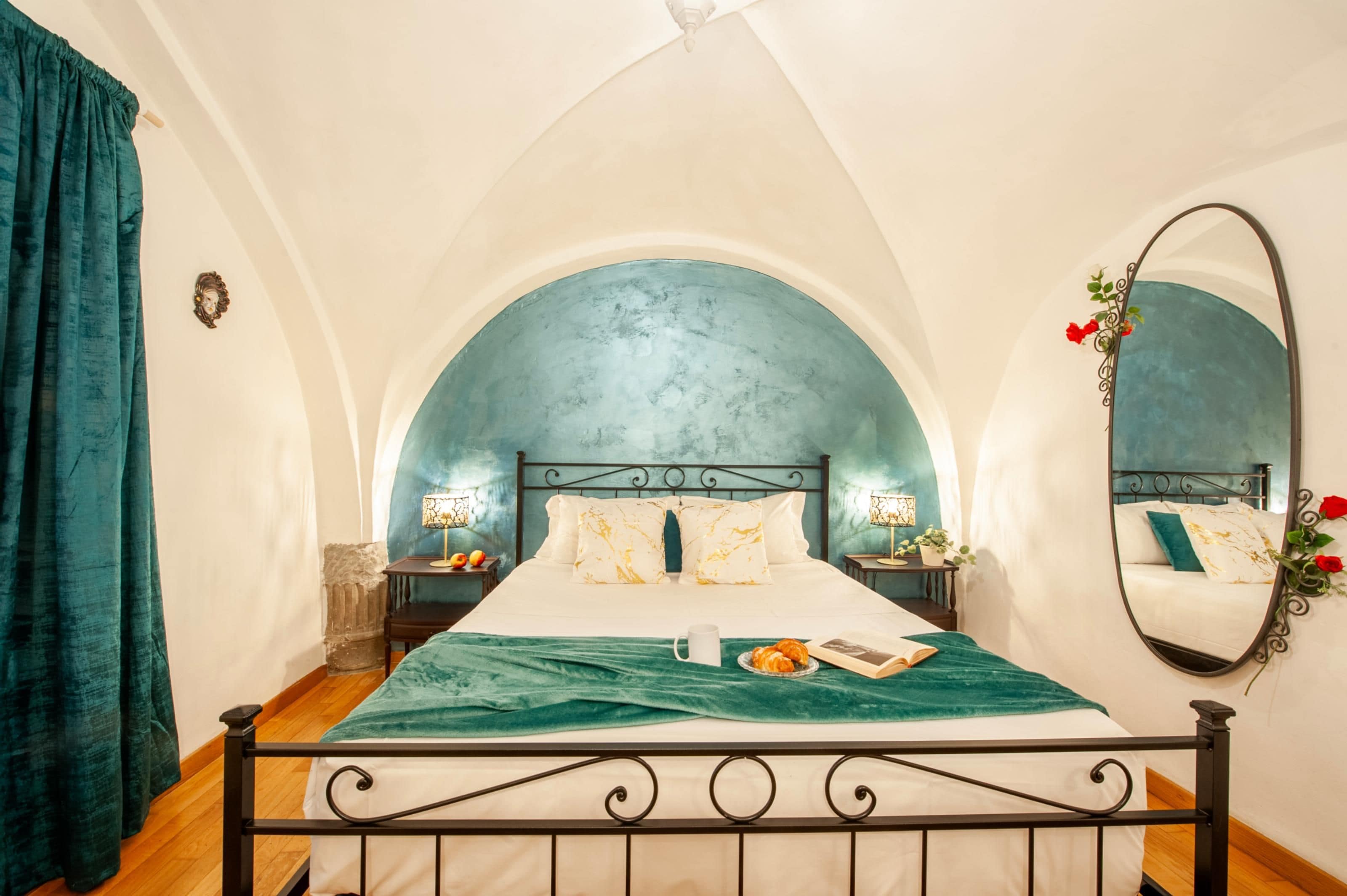A bedroom with a bed, a nightstand, a mirror, and a view of the sky through an arched window.