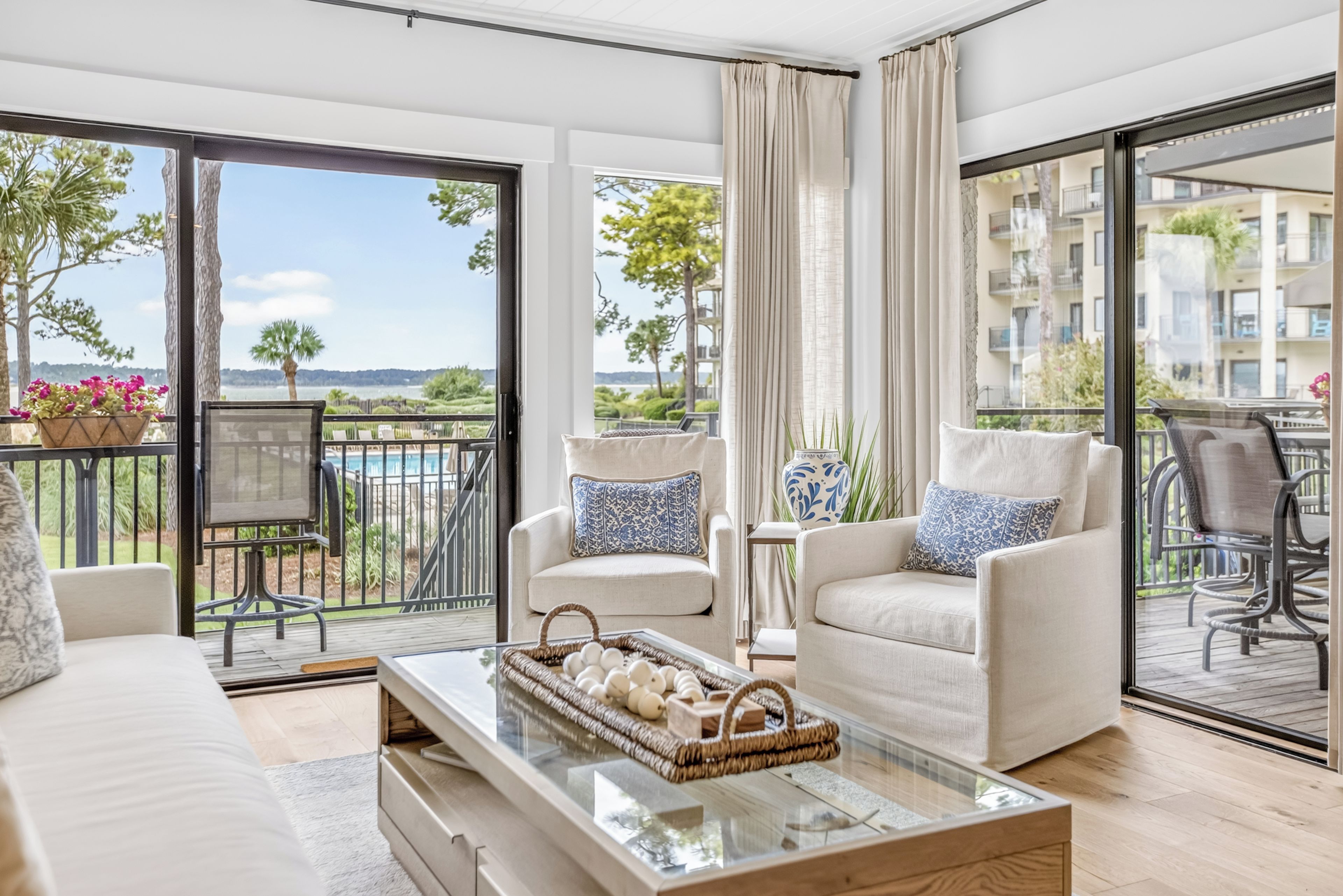 A modern living room with a sofa, armchairs, and a coffee table. There is a balcony with a view of the sea and buildings.