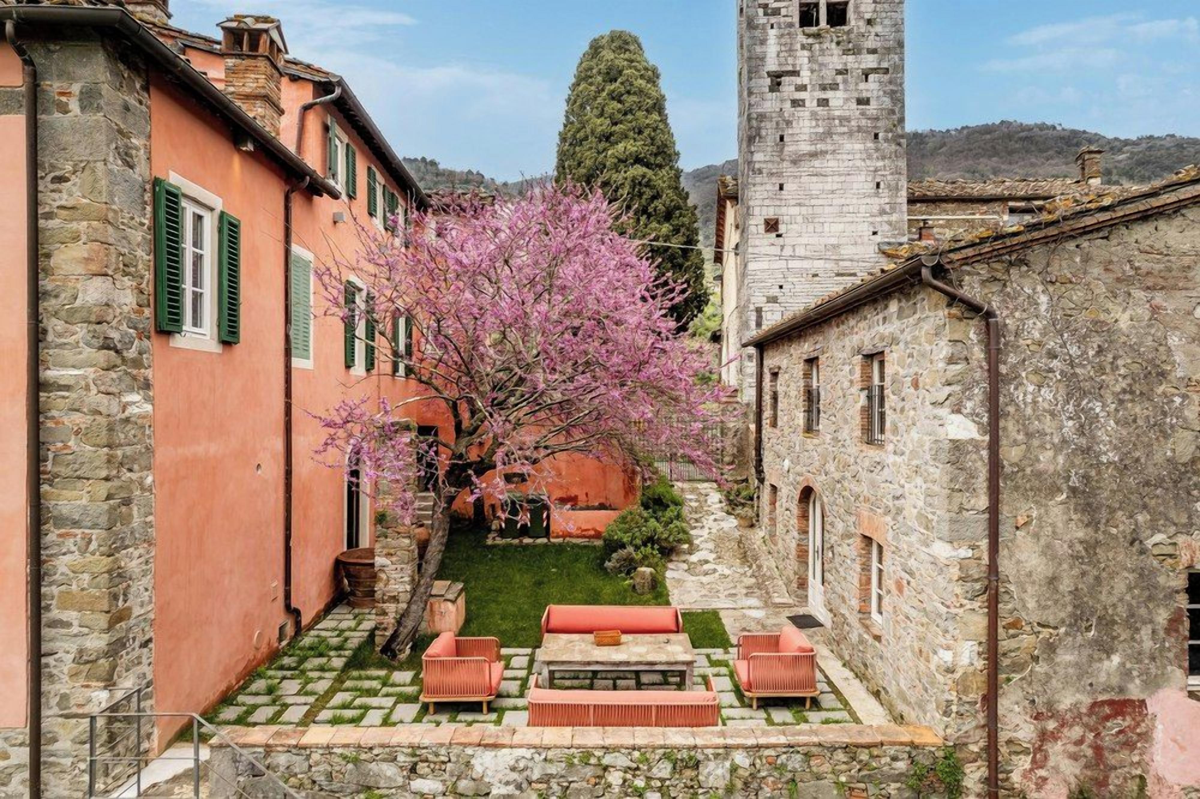 A courtyard with stone flooring, a pink flowering tree, and a stone tower in the background.