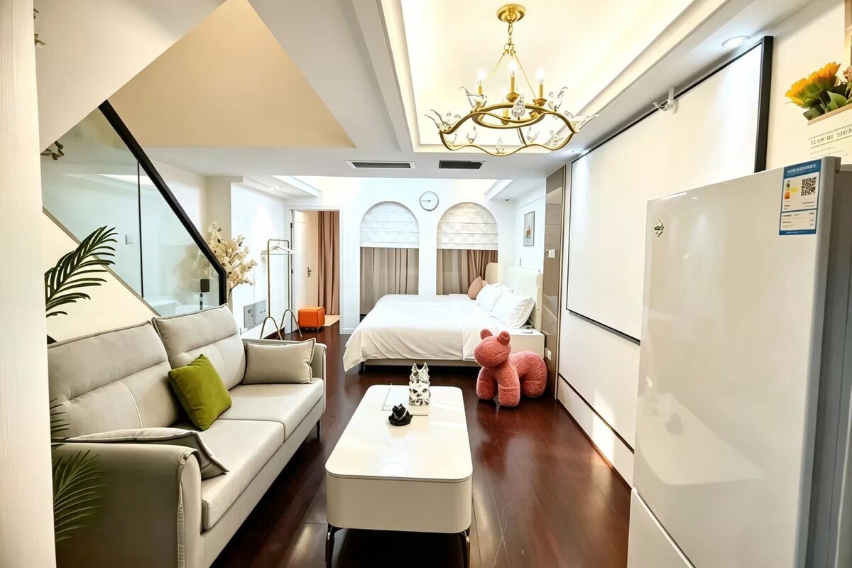 Quarto de hotel moderno com sofá, cama, mesa de centro, lustre e frigorífico.