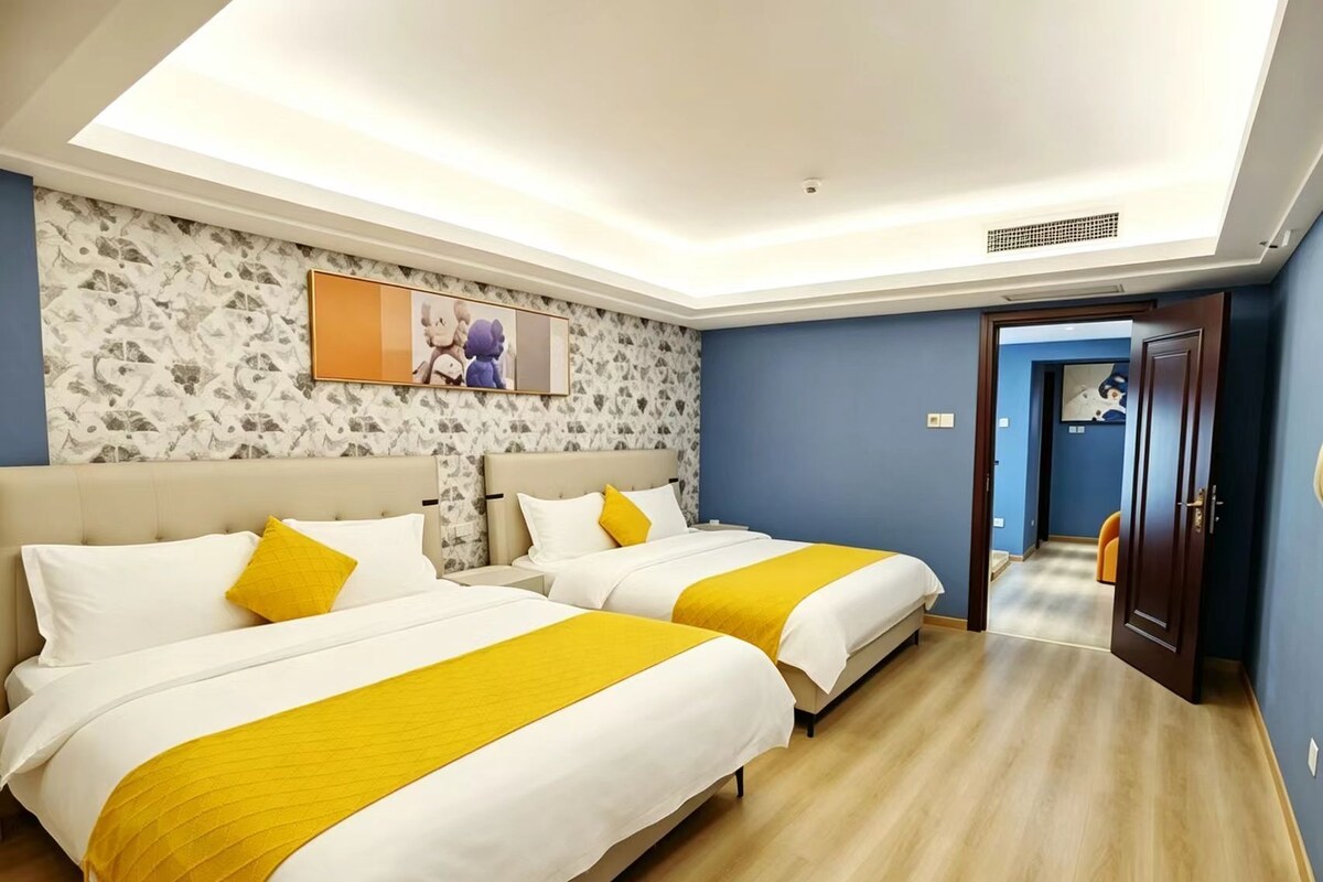 Quarto de hotel com duas camas, um claraboia, parede com padrão floral e porta que dá para outro cômodo.