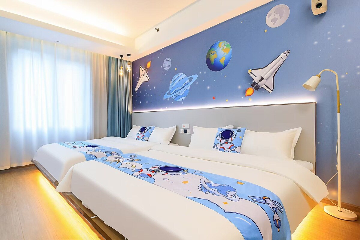 Quarto de hotel com uma cama grande, uma mesa de cabeceira, um abajur e uma parede decorada com tema espacial.