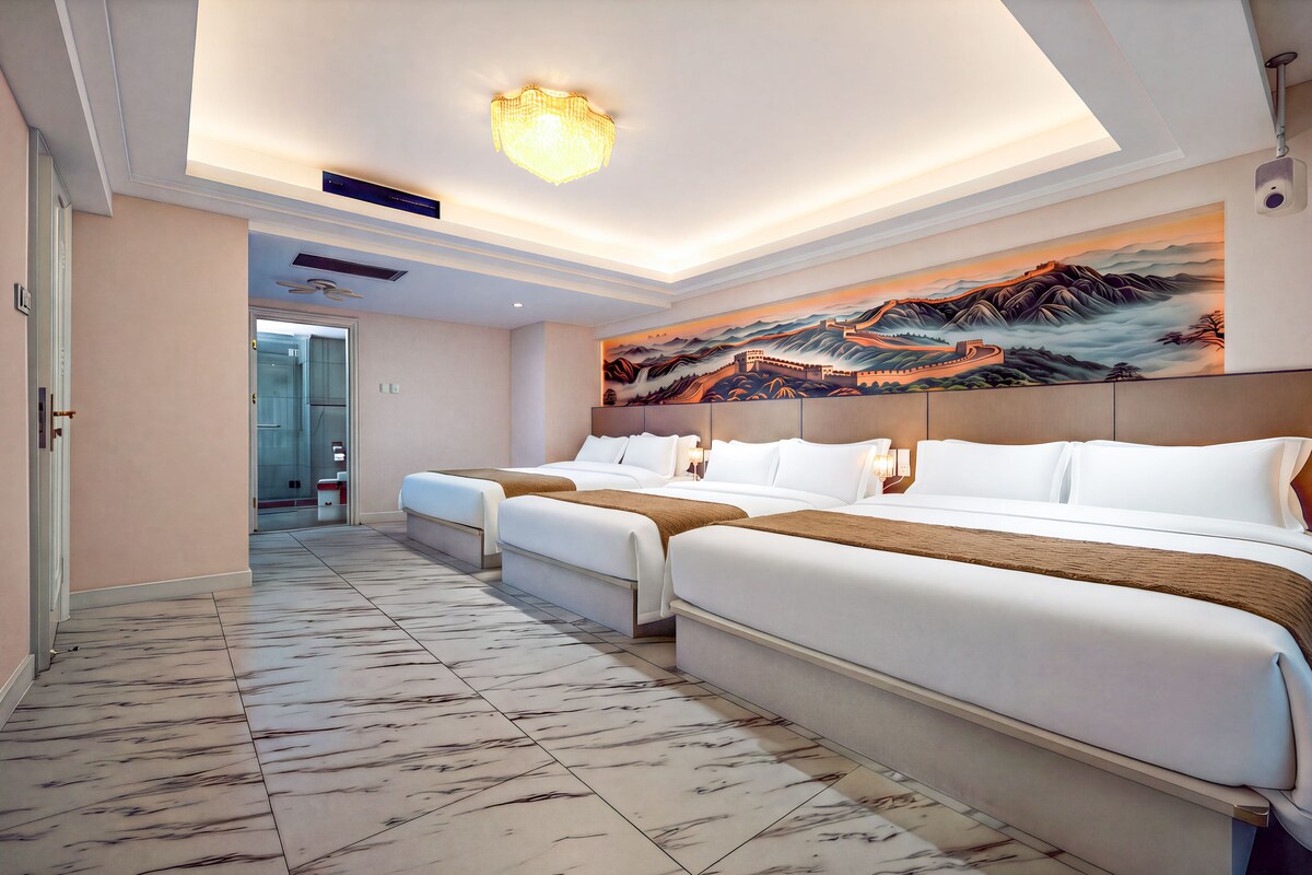 Quarto de hotel com uma cama grande, uma cama menor, um quadro de uma paisagem e um corredor que dá para um banheiro.