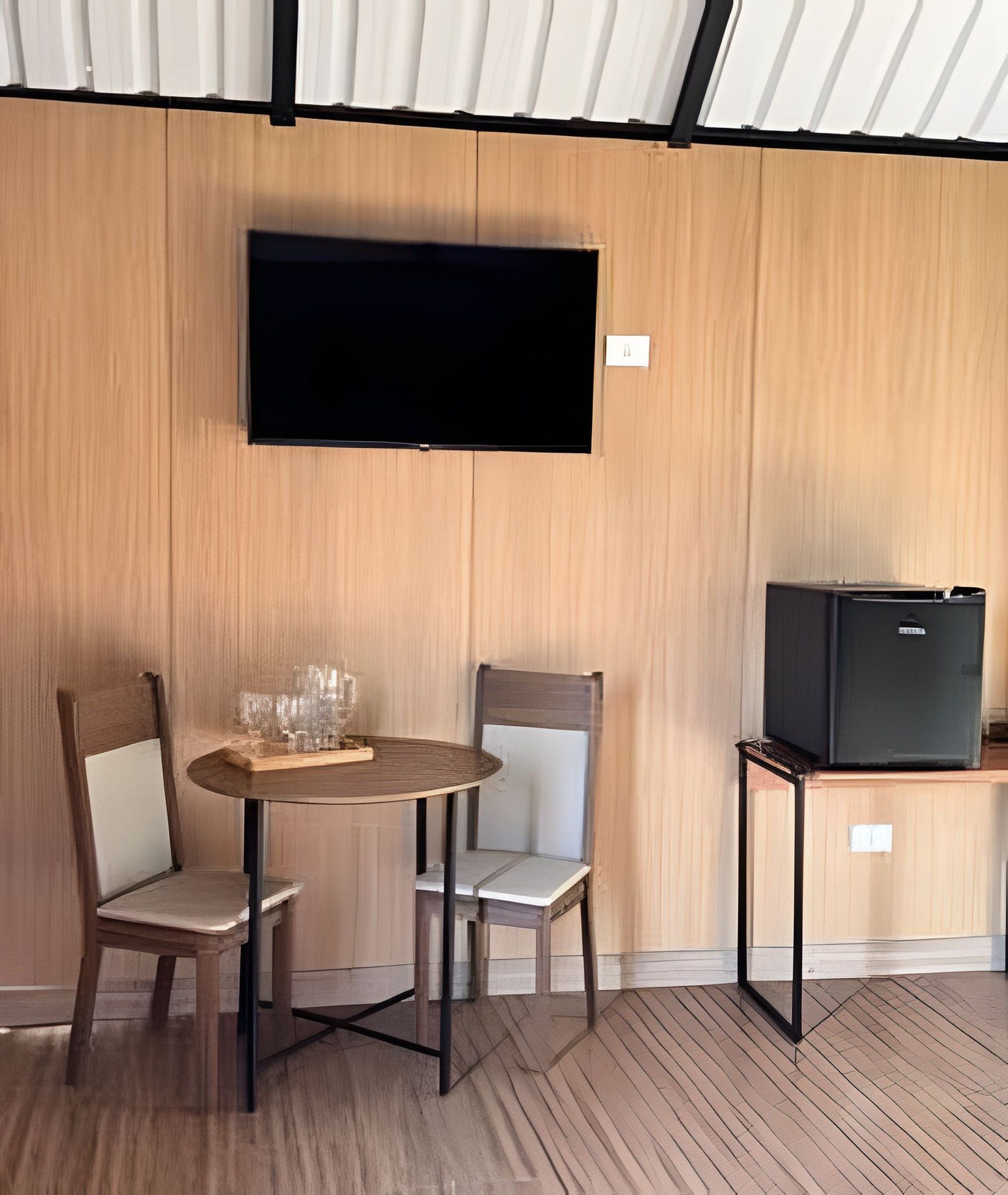 A room with a wooden wall, a flat-screen TV mounted above a table with two chairs, and a small black TV on a stand.
