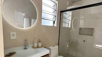 Casa de banho moderna com chuveiro envidraçado, espelho redondo e toucador com lavatório e porta-toalhas.