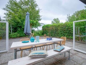 An outdoor patio with a table set for two, surrounded by wicker chairs, a bench, and a grey umbrella. - Holiday Home With 3 Bedrooms (De Cocksdorp)