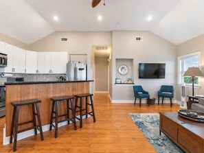 A modern kitchen with a wooden bar stool area, a dining area with a blue chair, and a living room with a flat-screen TV. - 7 Palms-3 bed/3 bath waterfront escape w/boat ramp (Sargent)