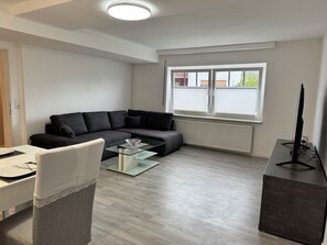 A modern living room with a sectional sofa, a glass coffee table, a flat-screen TV, and a window with blinds. - A Break in the Reinhardswald - Knickhagen Vacation Rental (Fuldatal)