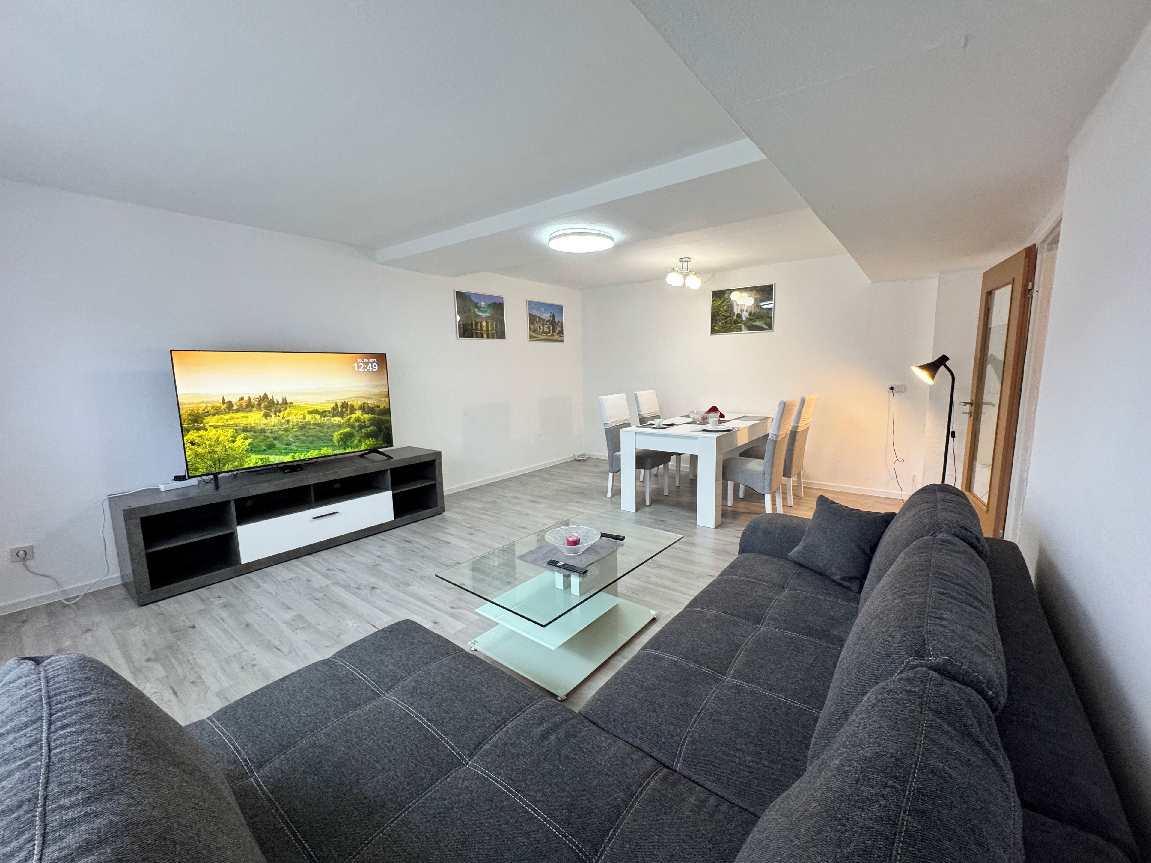 A modern living room with a flat-screen TV, a sofa, a glass coffee table, and a dining area with a white table and chairs.