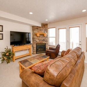 A living room with a stone fireplace, a television on a wooden stand, a leather sofa, and a balcony with white shutters. - Ski Condo and Summer Resort (Eden)
