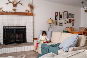 A cozy living room with a fireplace, a child reading, and a dining area with chairs and a table. - Walk to Downtown Lake Placid: 3BR Condo, Fireplace & Woodland Views (Lake Placid)