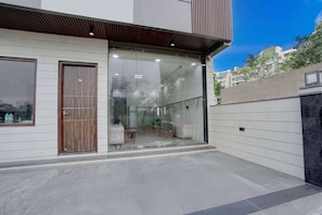 Un edificio moderno con un amplio acceso por puerta de vidrio, una puerta de madera a la izquierda y un muro de concreto a la derecha.