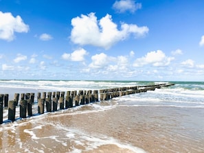 Ein Strand mit einem hölzernen Wellenbrecher, der sich ins Meer erstreckt, unter einem wolkenlosen Himmel.